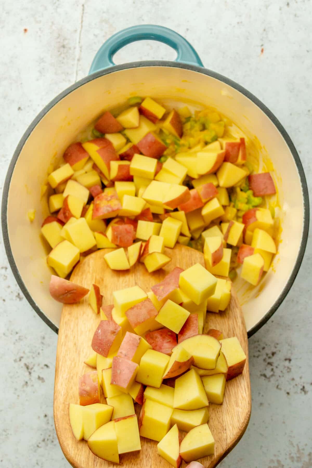 Cubes of potato are tossed into a soup on a Dutch oven on a light gray surface.