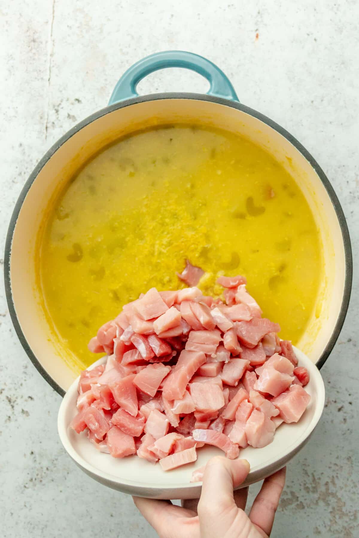 Cubes of thick ham are tossed into a soup in a Dutch oven on a light gray surface.
