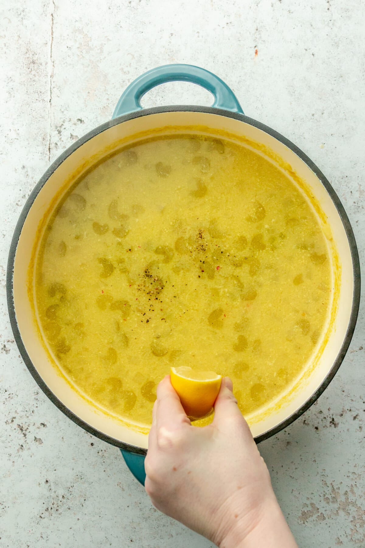 Lemon juice is squeezed into a soup in a Dutch oven on a light gray surface.