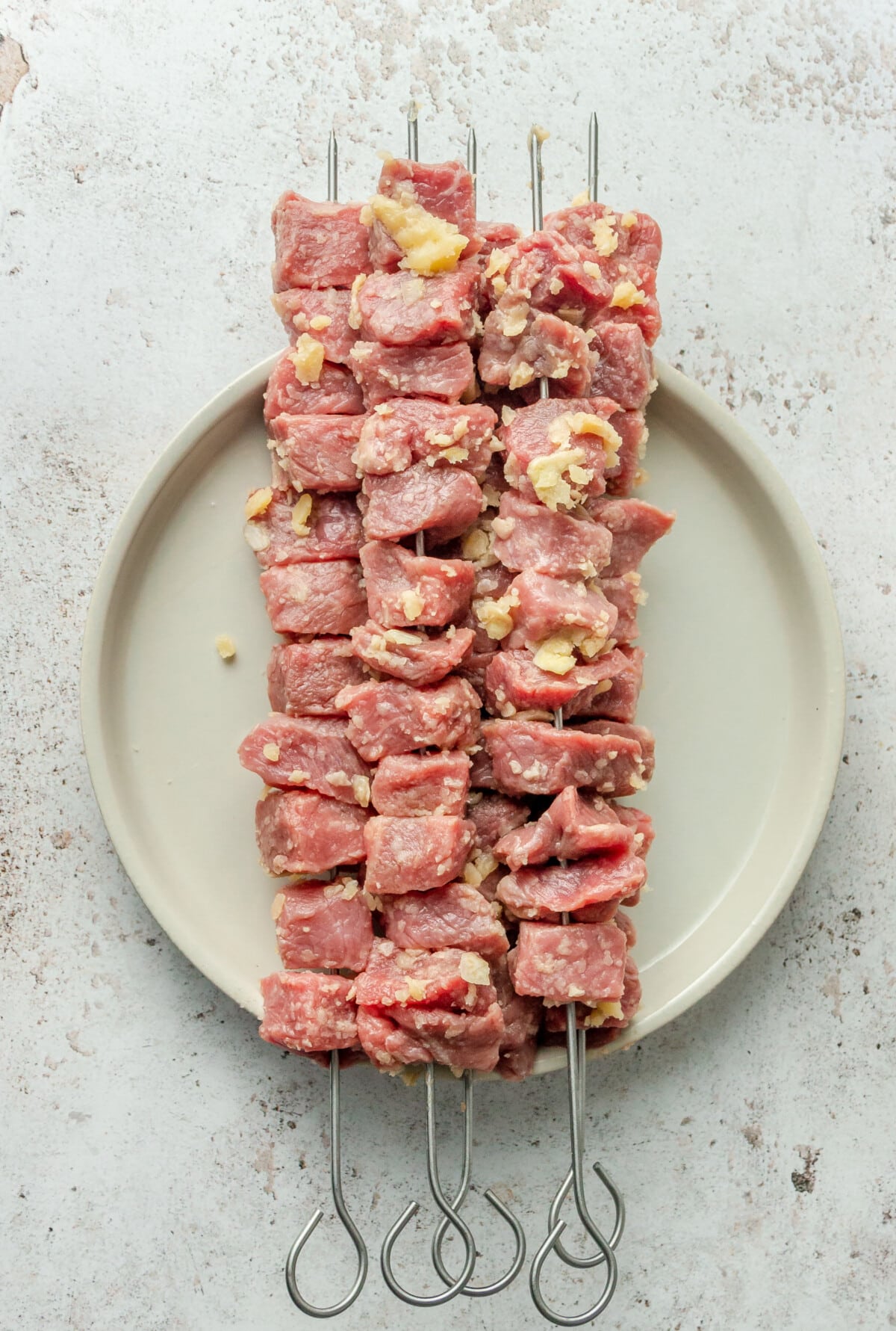 Cubes of steak sit on metal skewers on a plate ready for the grill on a light grey surface.