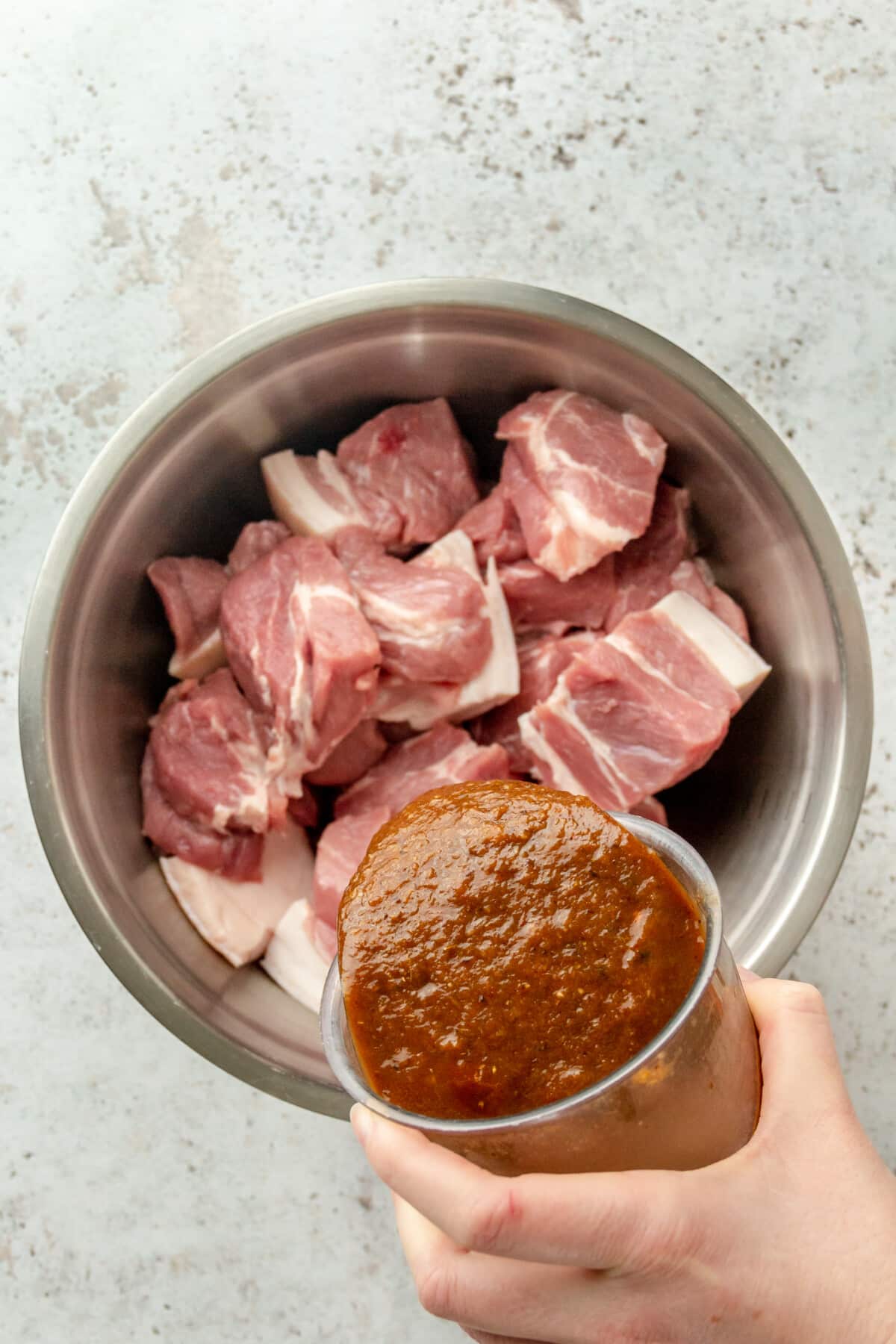 A marinade is poured over chunks of pork shoulder sitting in a stainless steel bowl on a light grey surface.