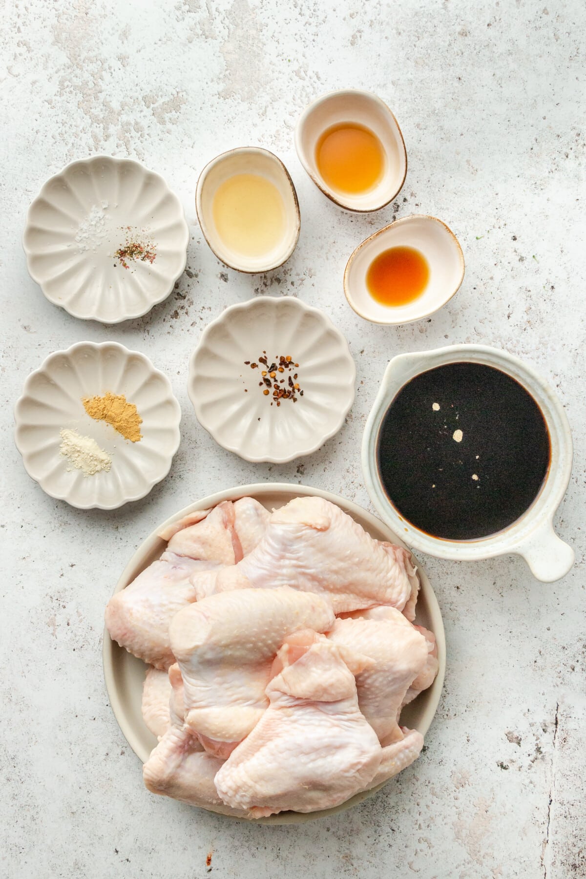 Ingredients for teriyaki chicken wings sit in a variety of plates on a light grey surface.