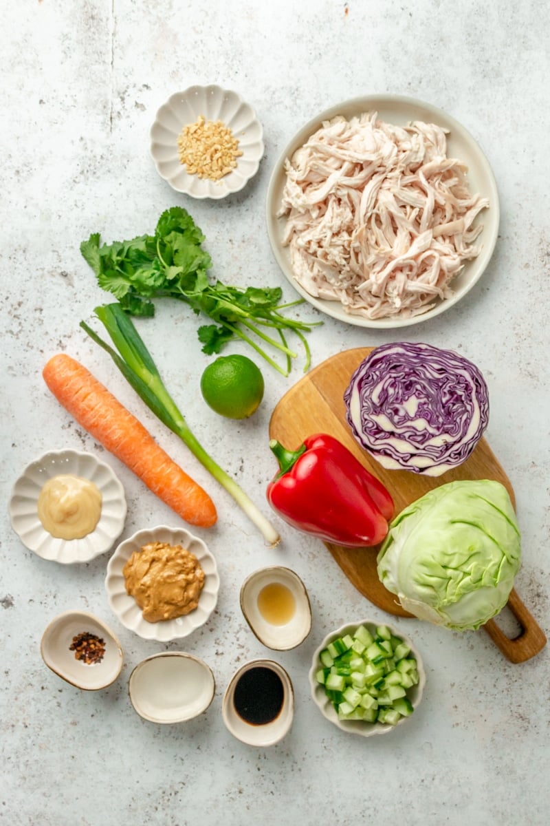 Ingredients for Thai inspired chopped chicken salad sit in a variety of bowl on a light grey countertop.