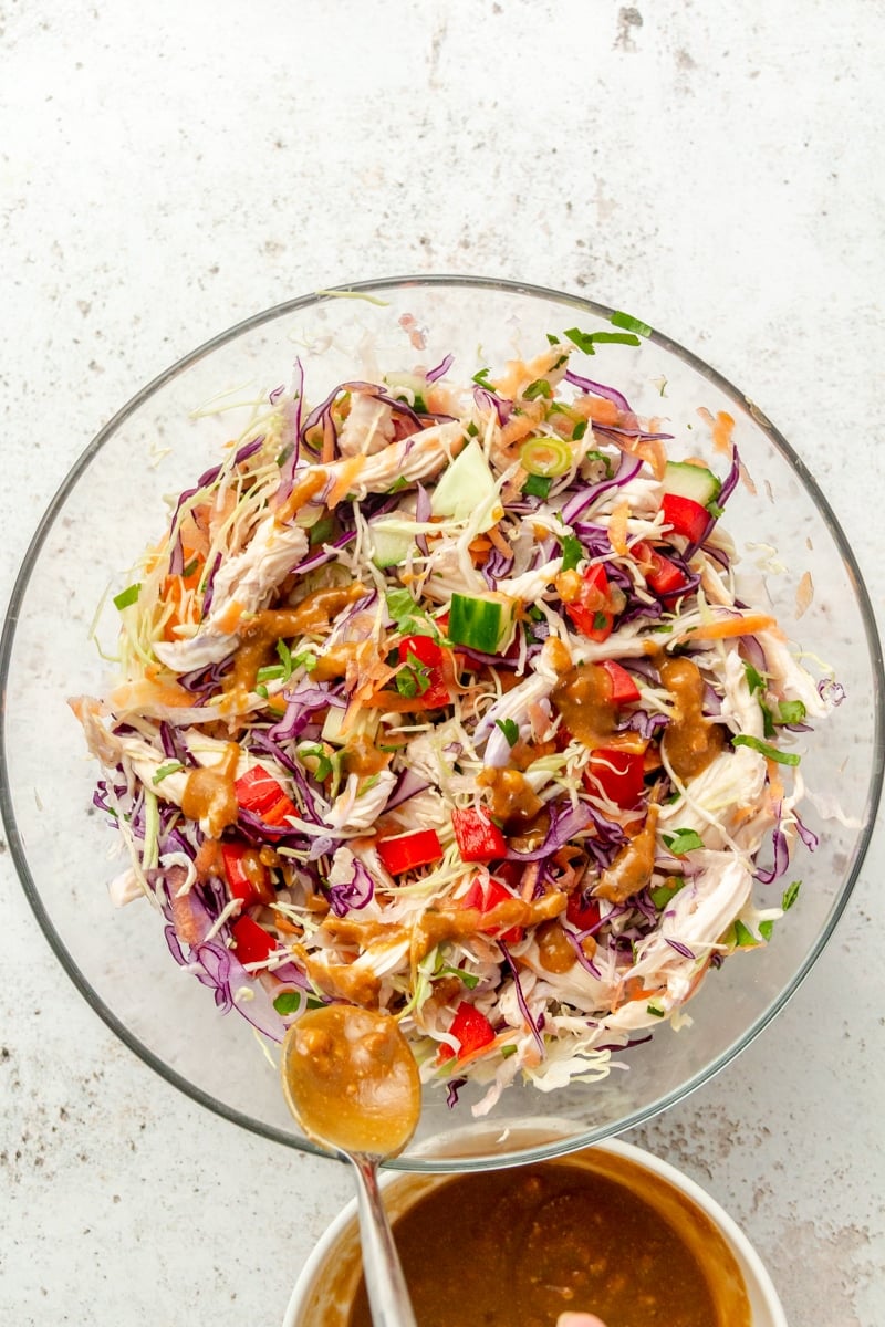 Thai inspired chopped chicken salad sits in a large glass mixing bowl. A brown sauce is shown being drizzled over the top.