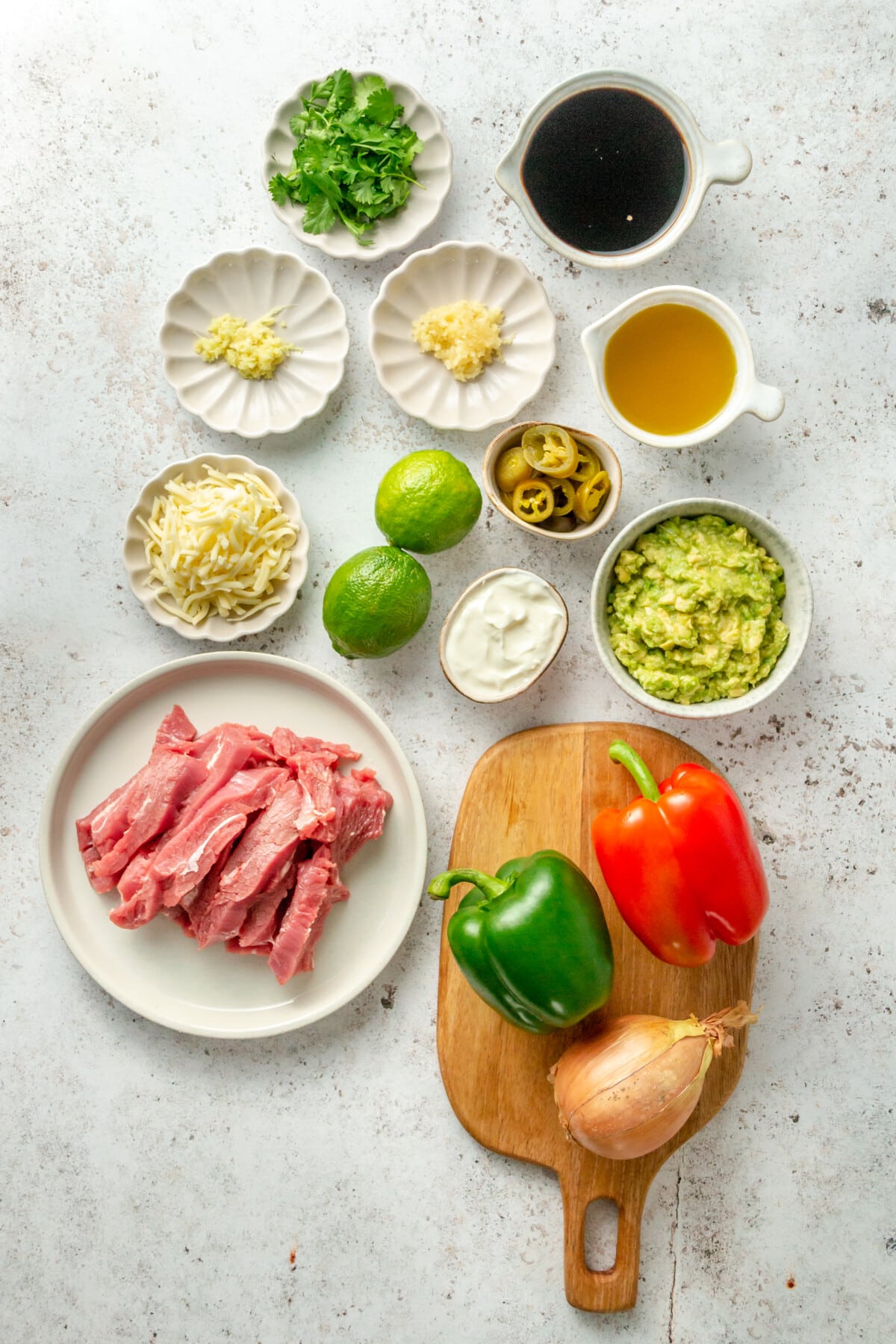 Ingredients for beef fajitas sit in a variety of plates and bowls on a light grey surface.