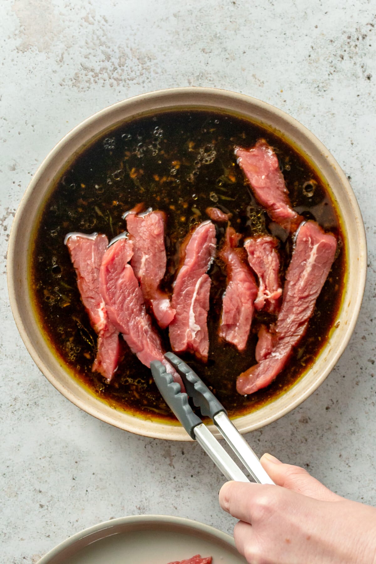 Beef strips are placed into a marinade in a shallow ceramic bowl on a light grey surface.