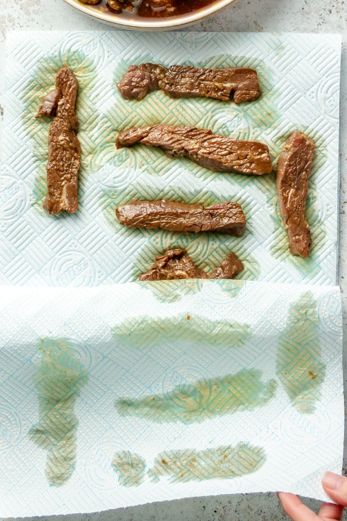 Marinated beef strips are dried in paper towels on a light grey surface.