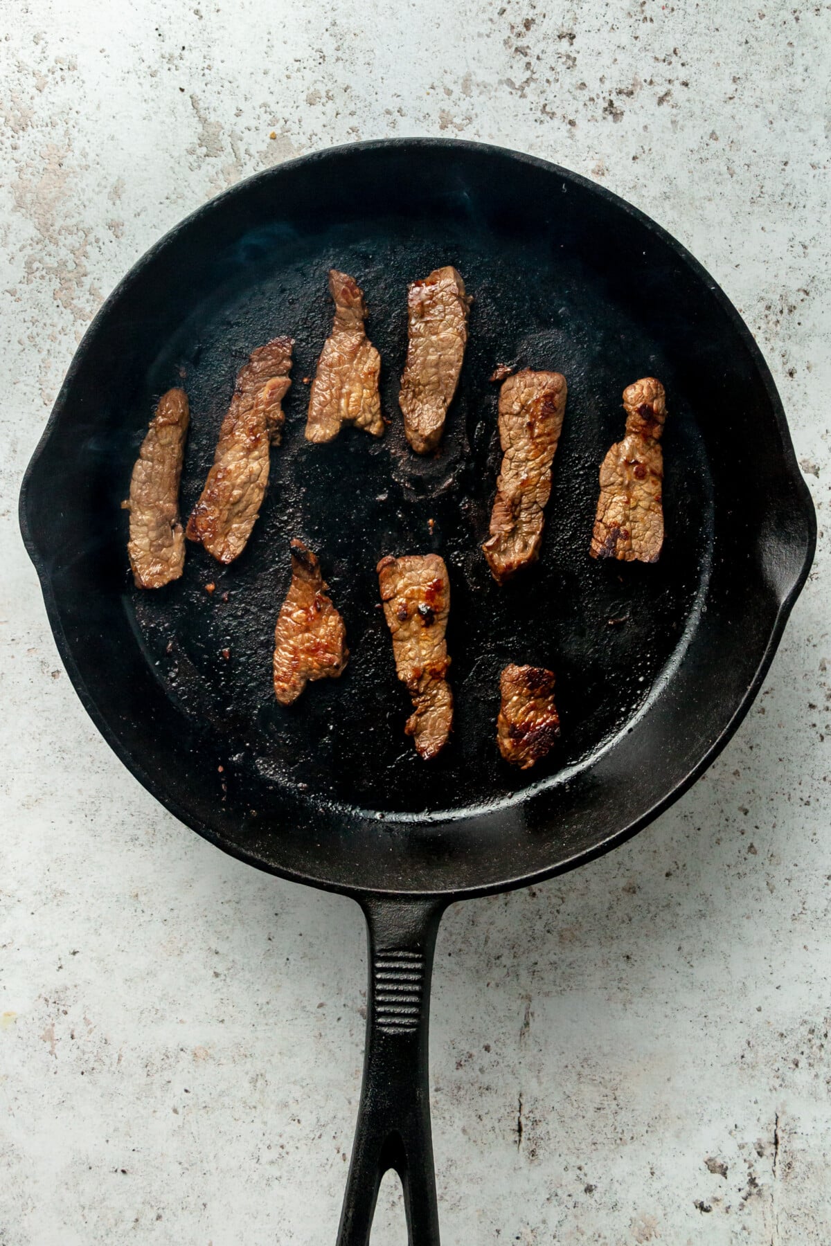Marinated beef strips sit in a cast iron skillet on a light grey surface.