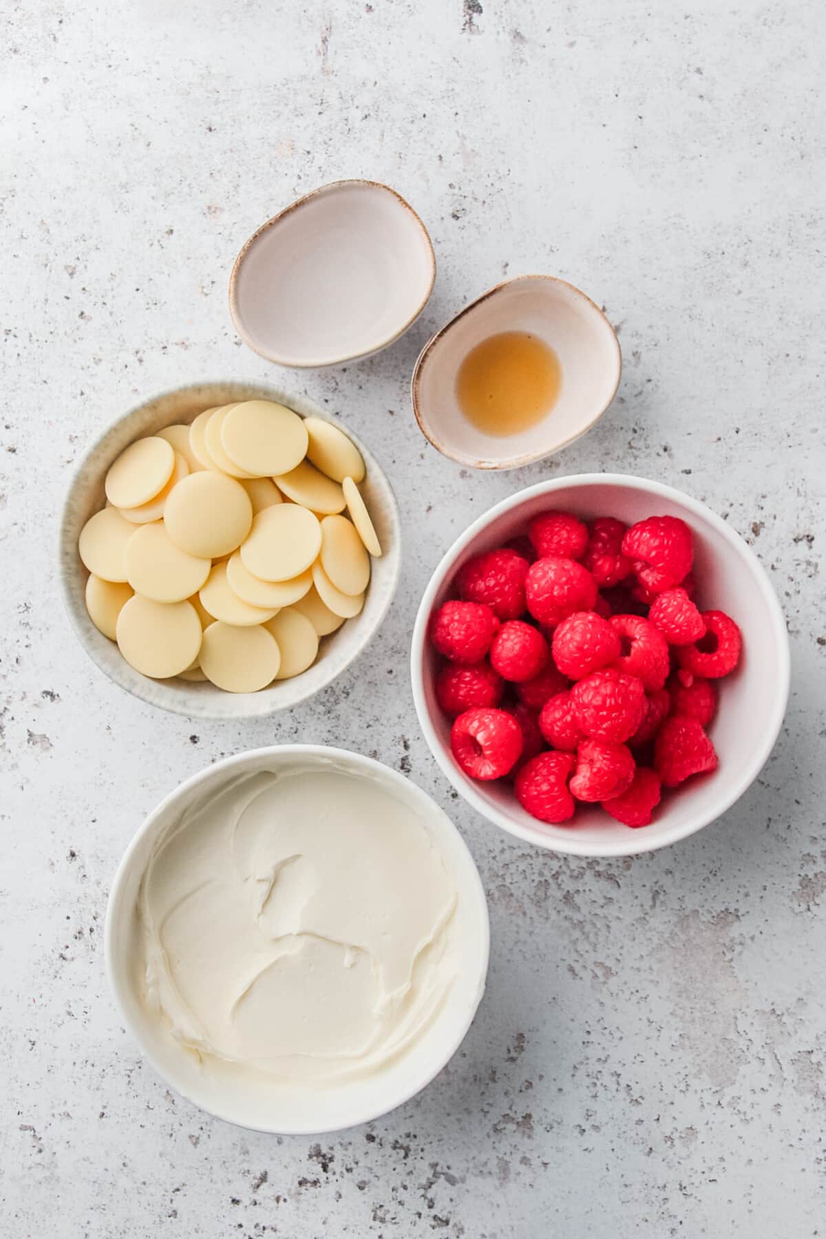 Ingredients for white chocolate raspberry cheesecake fat bombs are laid out in a variety of small plates and bowls on a light grey colored surface.