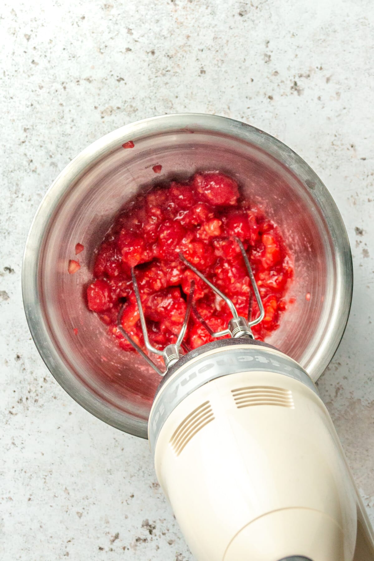 Fresh raspberries are whipped with a beater in a stainless steel bowl until muddled on a light grey colored surface.