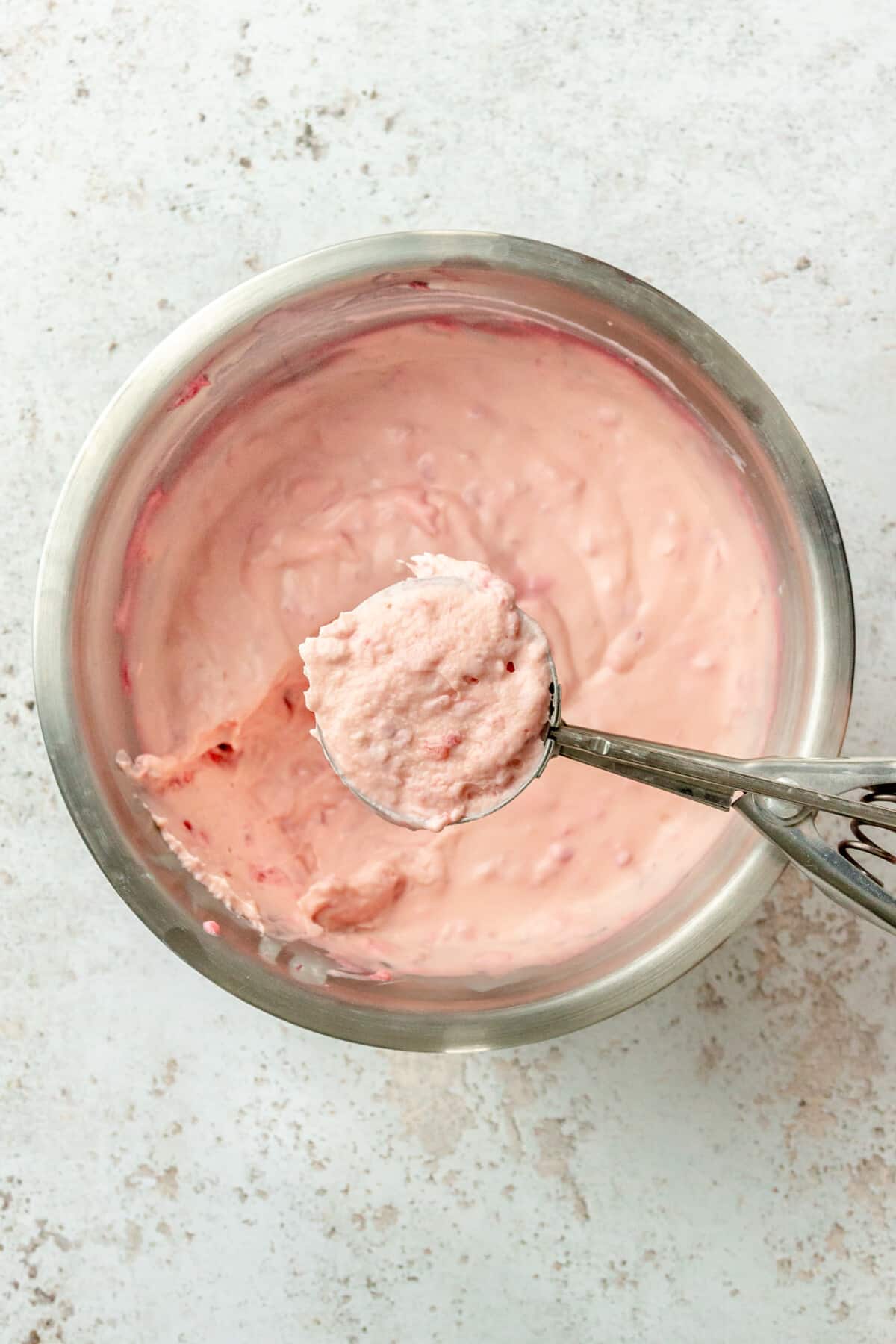 A small ice cream scoops up a chilled portion of the white chocolate raspberry cheesecake fat bombs from a stainless steel bowl on a light grey colored surface.