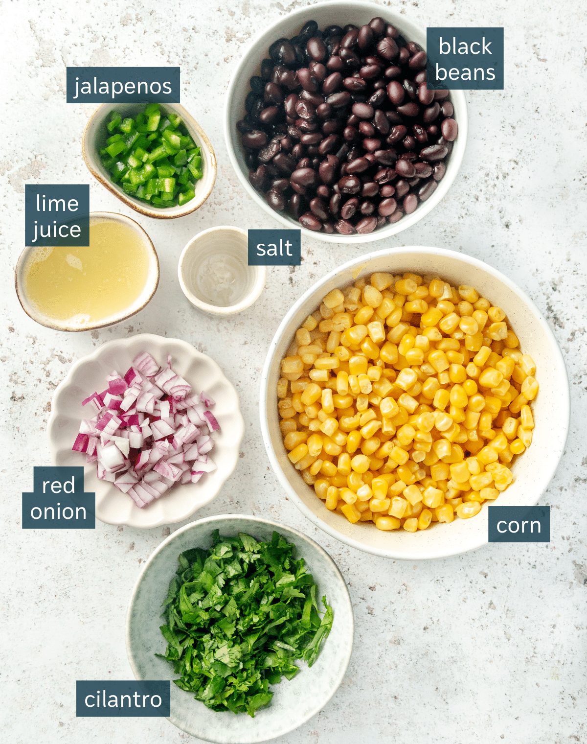 All of the ingredients needed for black bean and corn salsa portioned in different sized bowls on a light gray surface.