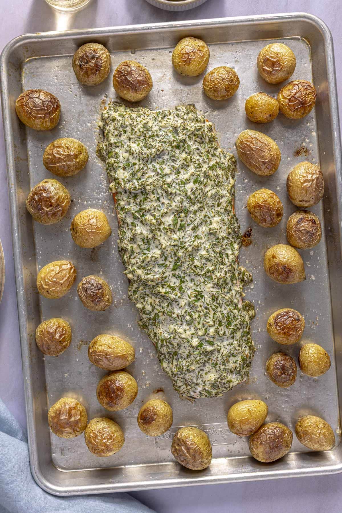A large salmon fillet covered with an herb mayo sauce on a sheet pan surrounded by roasted baby potatoes.
