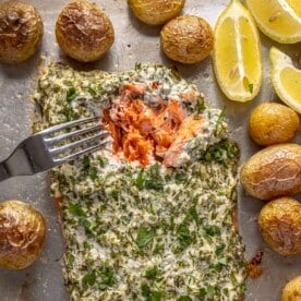 A large salmon fillet covered with an herb mayo sauce on a sheet pan surrounded by roasted baby potatoes.
