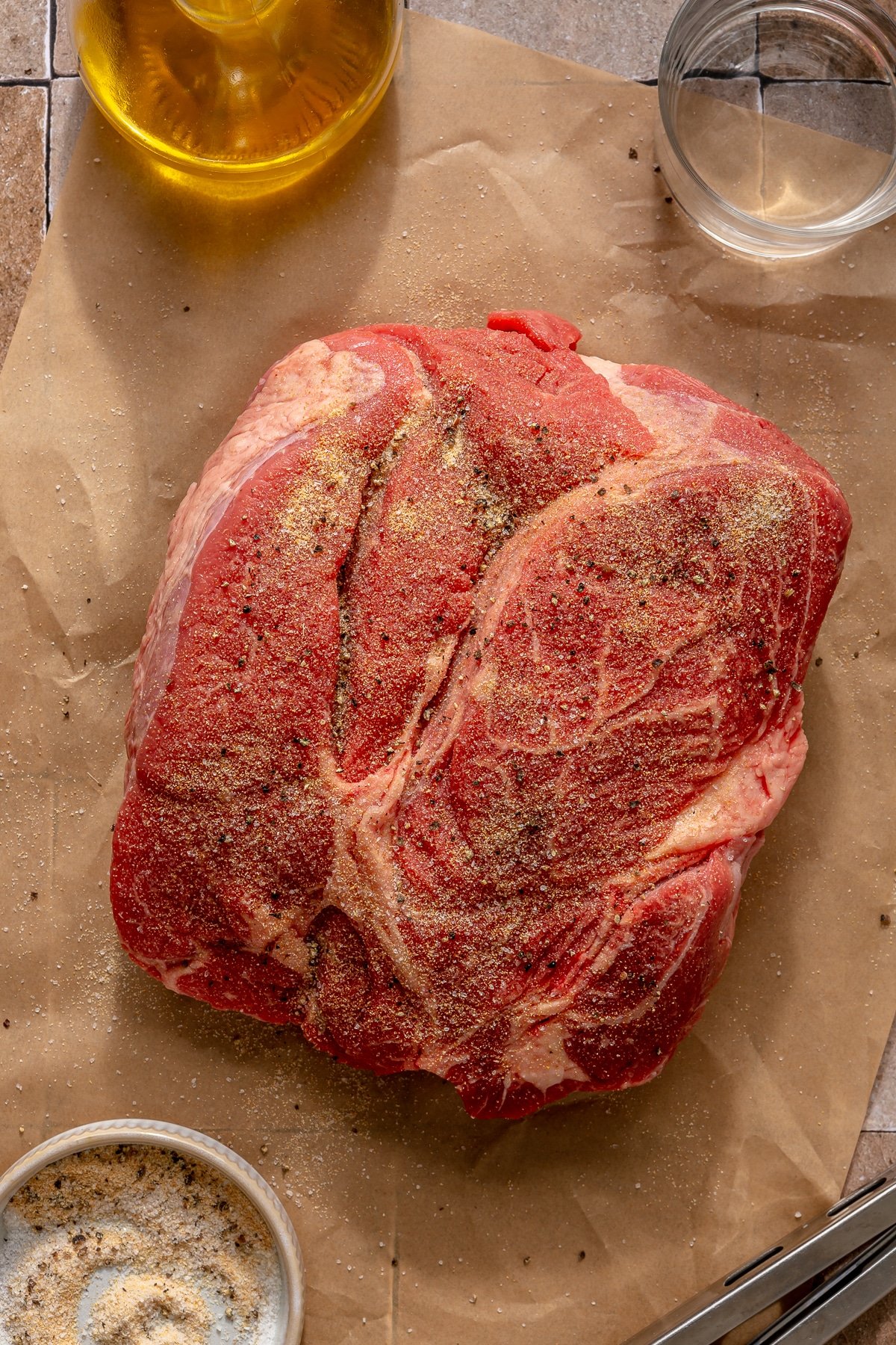 A large, seasoned chuck roast on parchment paper alongside other ingredients needed for Instant Pot pot roast.