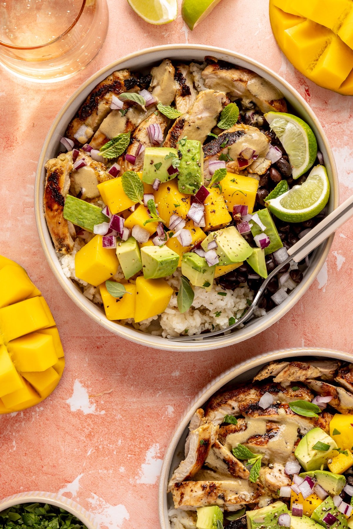 Mojo chicken bowls loaded with rice, beans, chicken, fresh mango, avocado, mint leaves, oregano leaves, and lime wedges.