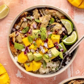 Mojo chicken bowls loaded with rice, beans, chicken, fresh mango, avocado, mint leaves, oregano leaves, and lime wedges.