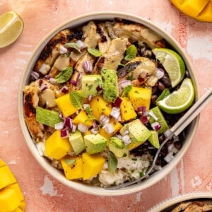 Mojo chicken bowls loaded with rice, beans, chicken, fresh mango, avocado, mint leaves, oregano leaves, and lime wedges.