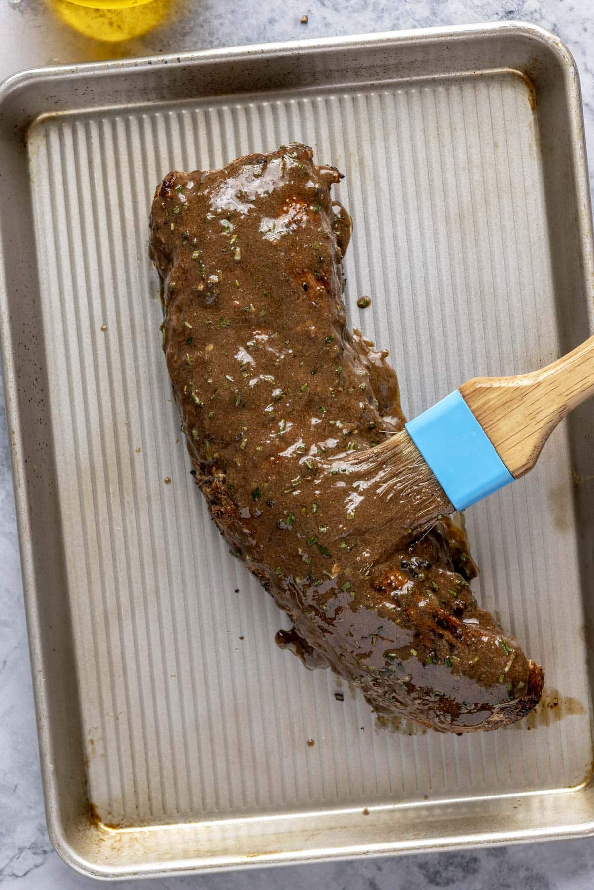 A person using a brush to baste a seared pork tenderloin on a sheet pan before going into the oven.