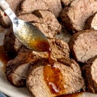 A platter of sliced pork tenderloin with someone drizzling sauce over one of the slices with a spoon.