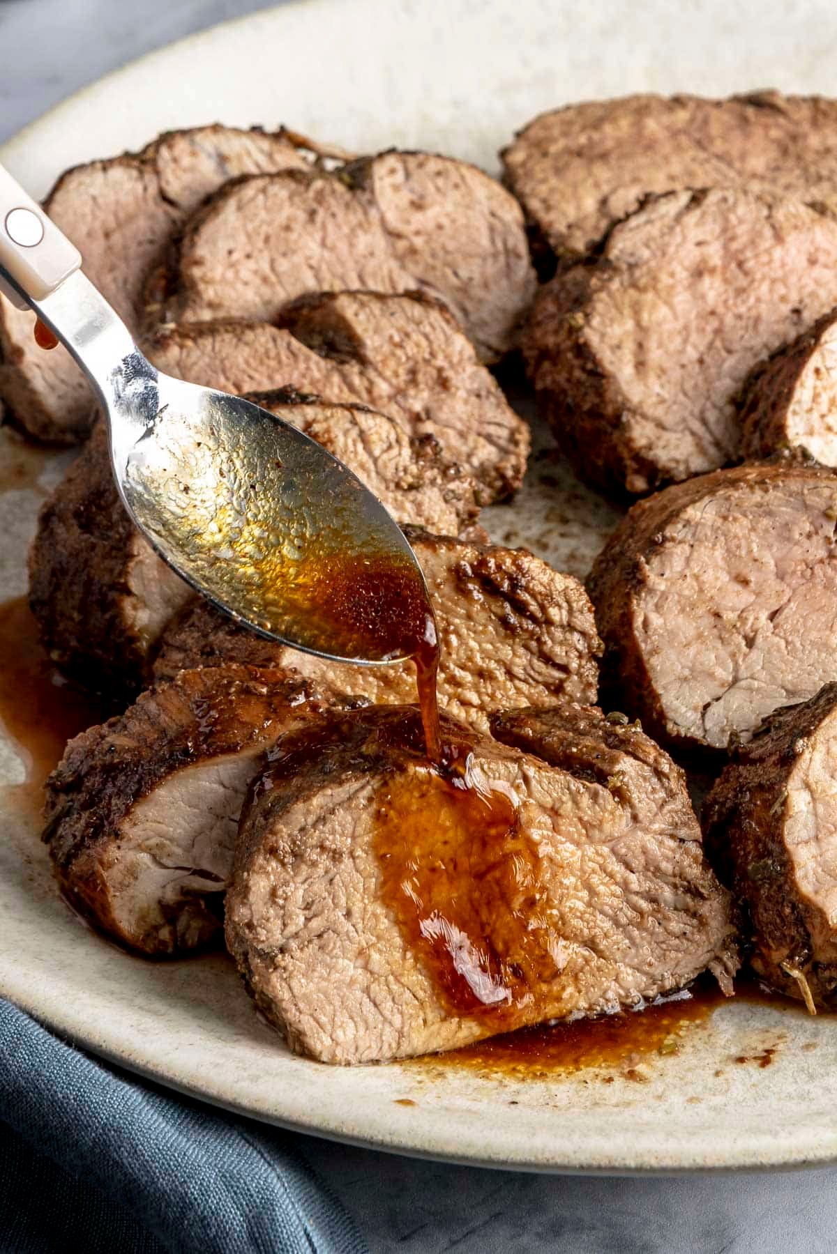 A platter of sliced pork tenderloin with someone drizzling sauce over one of the slices with a spoon.