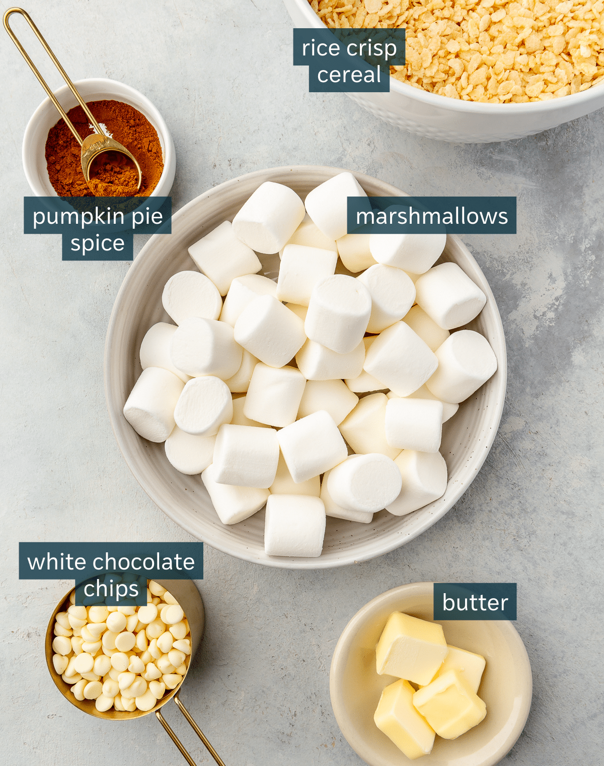 All of the ingredients needed for pumpkin spice rice krispie treats in different sized vessels on a gray surface.