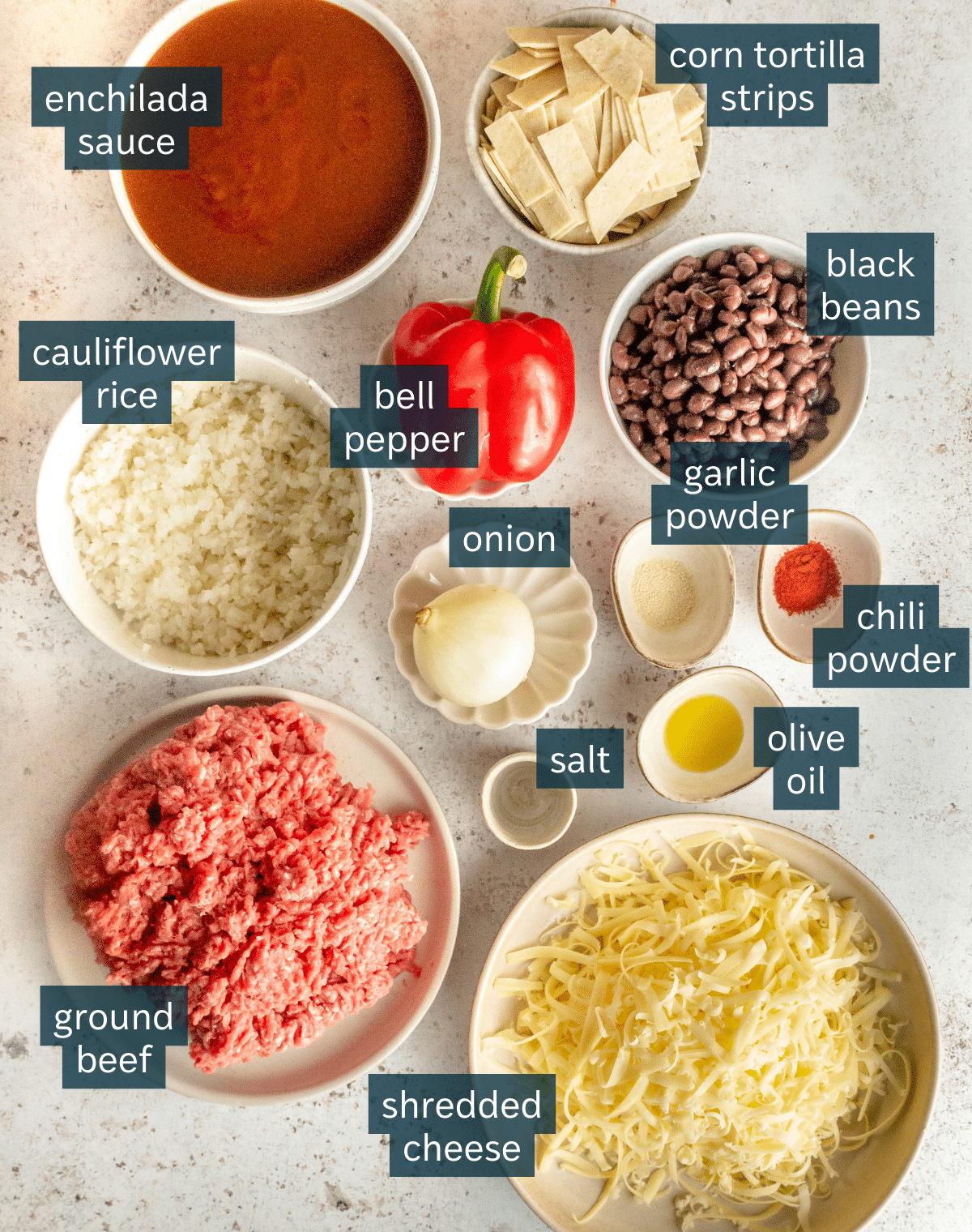 All of the ingredients needed for ground beef skillet enchiladas portioned out in different sized bowls and plates on a light gray surface.