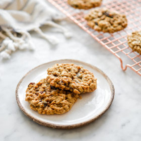 Two oatmeal raisin cookies sitting on a white plate with brown edge and copper cooling rack with cookies in the background.