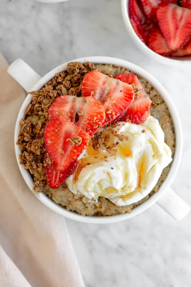 Top down view of oatmeal in a white mug topped with sliced strawberries, granola, mascarpone, and honey. 