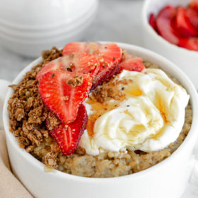 White bowl with oatmeal topped with sliced strawberries, granola, mascarpone, and a drizzle of honey.