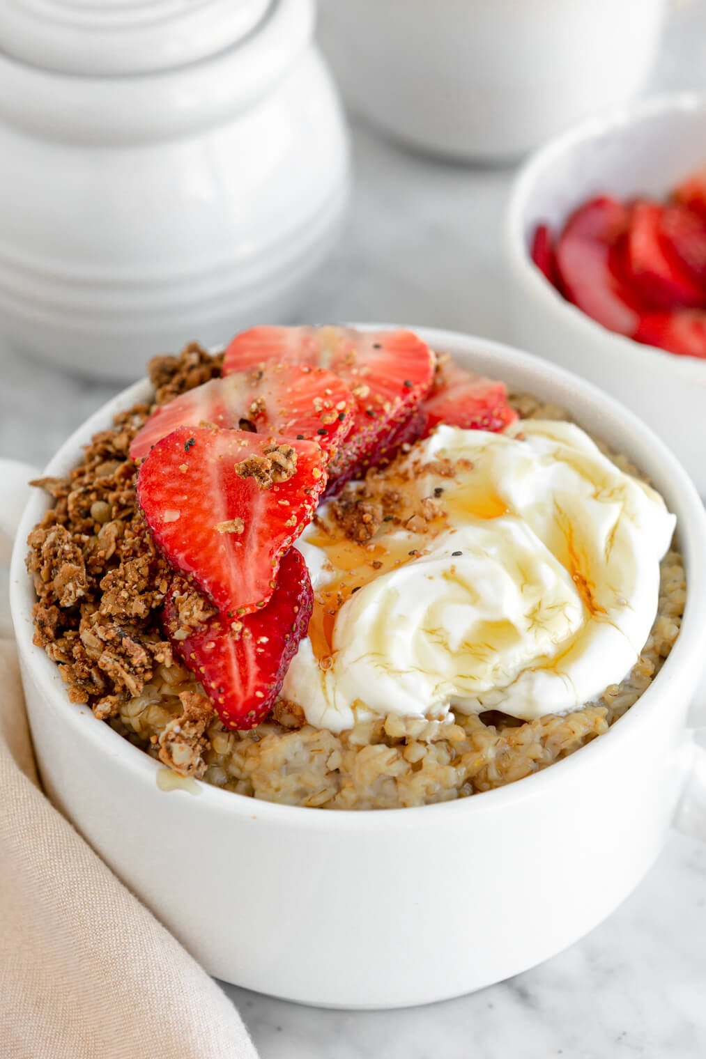 White bowl with oatmeal topped with sliced strawberries, granola, mascarpone, and a drizzle of honey.