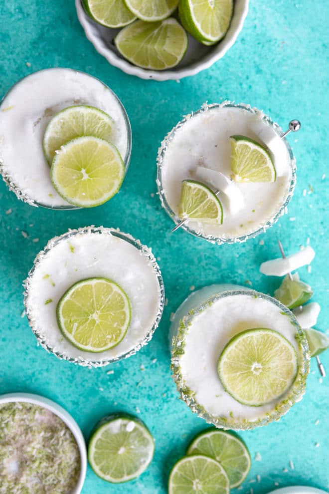 Top down view of 4 coconut margaritas with various lime and coconut garnishes on a teal background.