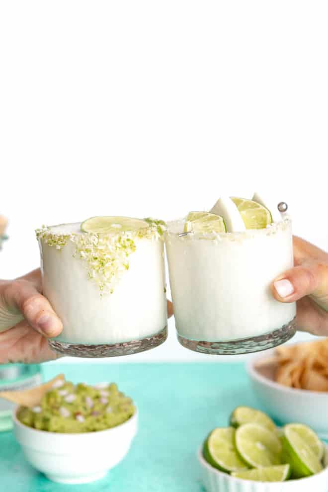 Two hands holding coconut margarita glasses together with guacamole, bowl of limes, and chips in the background.