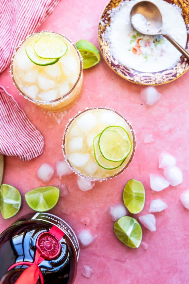 Top down view of Grand Marnier margaritas garnished with 2 slices of limes, a Grand Marnier Bottle, and a saucer with sea salt with a copper spoon on a pink blush surface with squeezed lime wedges, sea salt flakes, and ice cubes on the table.