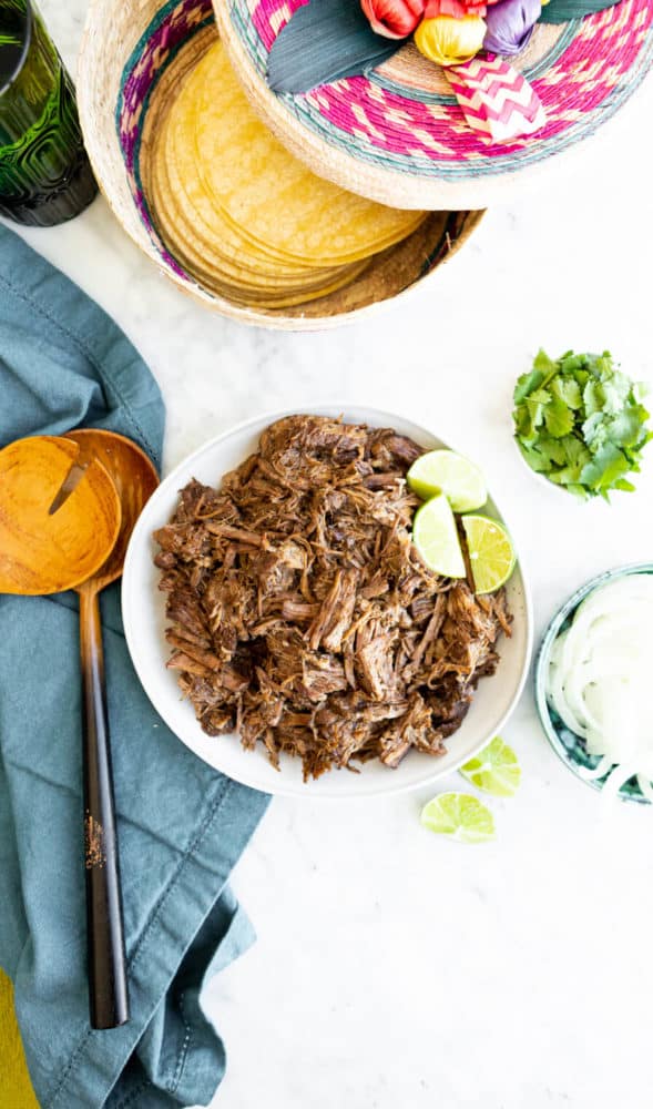 Top down view of white bowl with barbacoa beef garnished with 3 lime wedges surrounded on one side by a teal napkin and serving utensils and a bowl of cilantro and onions on the other and a basket of tortillas to the top. 