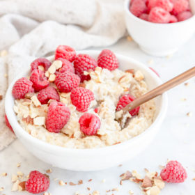 Oatmeal in a white bowl topped with chopped almonds and raspberries with a small white bowl of raspberries and a beige linen in the background.