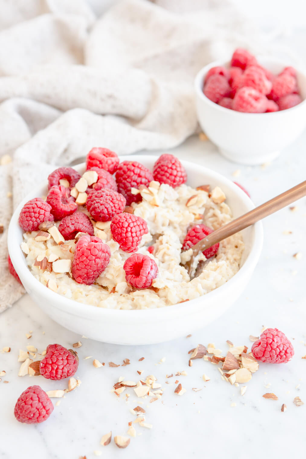 Oatmeal in a white bowl topped with chopped almonds and raspberries with a small white bowl of raspberries and a beige linen in the background.