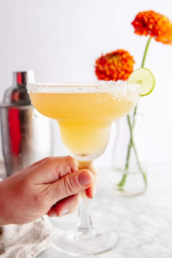 Hand holding classic margarita glass with Cadillac Margarita served neat with flaky sea salt rim and slice of lime on the side with shaker bottle and orange flowers in the background.