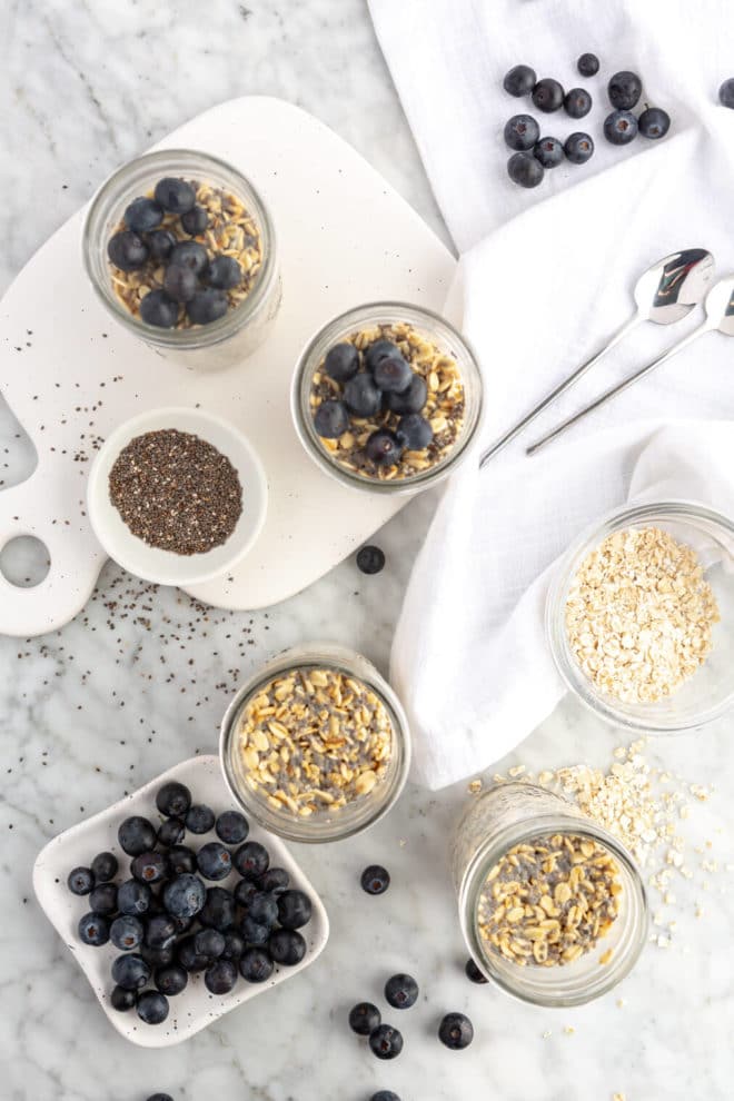 Top down view of gray and white countertop with fixings for overnight oats including plate with blueberries, small mason jars with overnight oats, chia seeds, spoons, linen napkins, and dry oats.