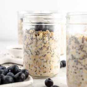 Small mason jar with overnight oats topped with blueberries next to a plate with blueberries and a sprinkle of blueberries on the counter next to it.