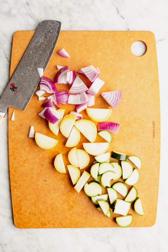 Chopped and sliced vegetables on a epicurean cutting board with a butcher knife on a marble surface.