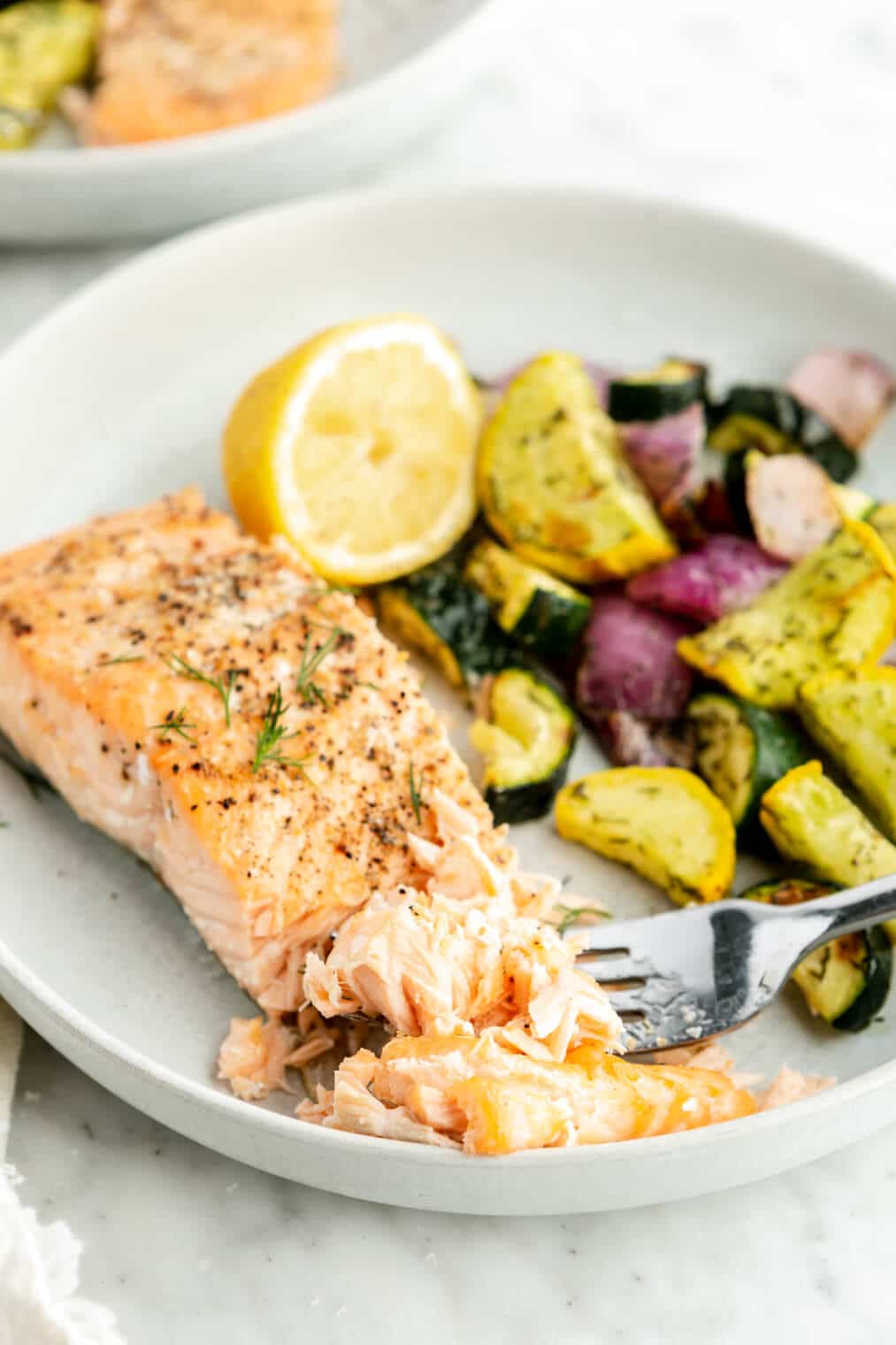 Cooked salmon filet on a plate with a fork flaking a bite of salmon. There is also a medley of vegetables on the plate.