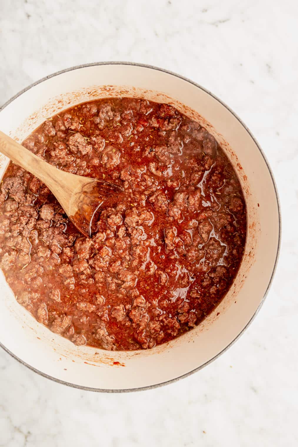 Ground beef in a tomato and wine sauce in a round, white casserole dish with a wooden spoon resting inside.