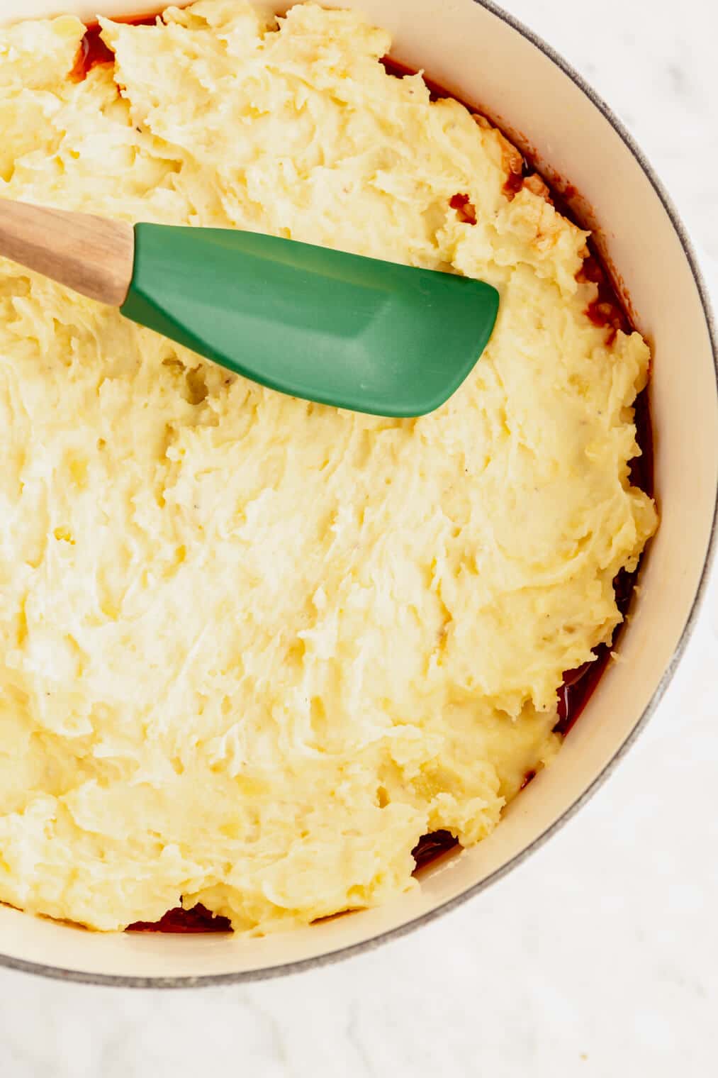 Green spatula spreading out mashed potatoes on top of beef mixture in a round, white casserole dish.