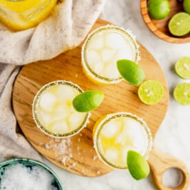 Top down view of three mezcal margaritas on a wooden cutting board, all garnished with a wedge of lime. On a table with a bowl of limes and peel container with salt, beige linen, and jar of margarita mix.