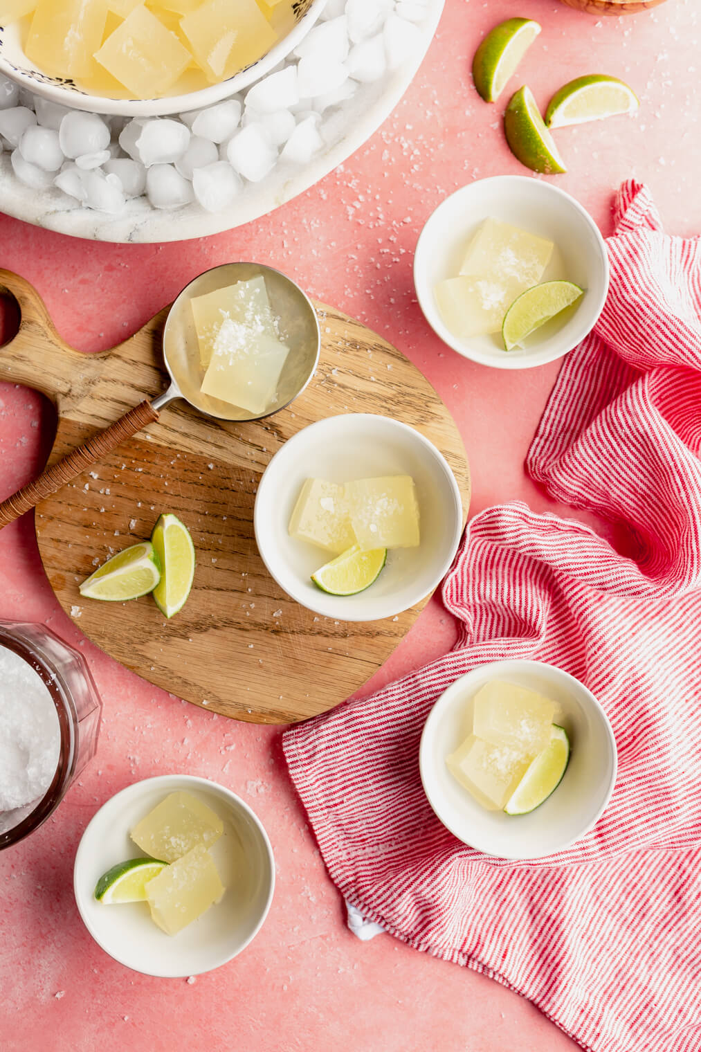 Top down view of margarita jello shots in small white bowls on a blush colored table draped with a red and white striped towel.