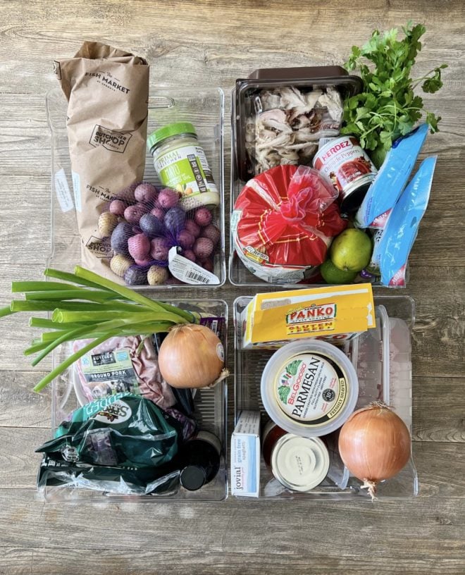 Top down view of 4 plastic container bins filled with groceries on a wooden surface.