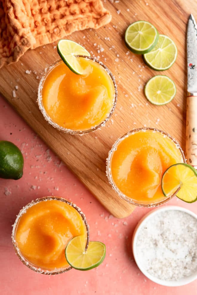 Top down view of frozen peach margaritas in glasses garnished with flaky sea salt and a slice of lime on a pink colored surface with a cutting board, paring knife, sprinkle of flaky sea salt, lime slices, and orange linen.