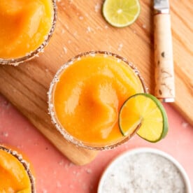 Top down view of frozen peach margaritas in glasses garnished with flaky sea salt and a slice of lime on a pink colored surface with a cutting board, paring knife, sprinkle of flaky sea salt, lime slices, and orange linen.