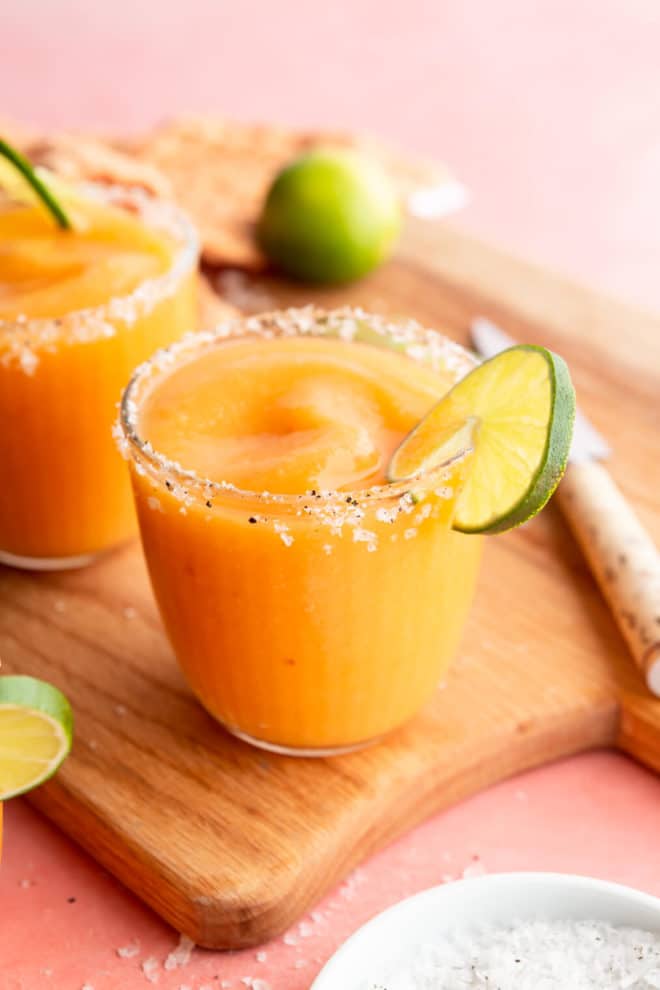 Side view of frozen peach margaritas in glasses garnished with flaky sea salt and a slice of lime on a pink colored surface with a cutting board, paring knife, sprinkle of flaky sea salt, lime slices, and orange linen.
